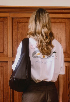 Femme aux cheveux blonds longs et ondulés portant un T-shirt blanc avec un motif d'œufs au plat et de café, portant un sac à bandoulière noir, debout devant un panneau en bois.