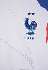 Jersey deportivo blanco con un emblema de gallo bordado en azul y "FFF" debajo. Incluye colores de acento en rojo y azul con patrones geométricos.