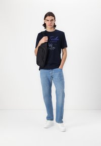 Marineblå t-skjorte med trykket tekst, lyseblå jeans, svart crossover-veske og hvite joggesko. Modellen står mot en hvit bakgrunn.
