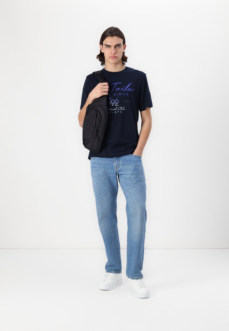 Marineblå t-skjorte med trykket tekst, lyseblå jeans, svart crossover-veske og hvite joggesko. Modellen står mot en hvit bakgrunn.