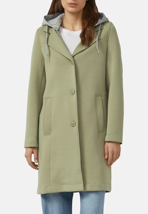 Frau trägt einen hellgrünen, geknöpften Mantel mit Seitentaschen und einer grauen Kapuze über einem weißen Oberteil und blauen Jeans.