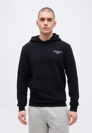 Mladý muž s krátkymi vlasmi, oblečený v čiernej mikine značky Björn Borg a svetlosivých teplákoch, stojaci pred jednoduchým svetlým pozadím.