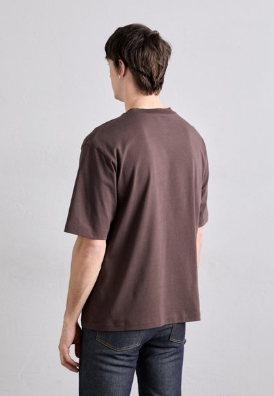 Camiseta de algodón marrón con mangas cortas, cuello redondo y un ajuste relajado, combinada con jeans oscuros. Diseño sencillo, textura suave, sin patrones.