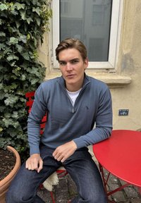 Jaunas vyras mėlynu megztiniu ir džinsais sėdi ant raudonos kėdės prie raudono apvalaus stalo lauke šalia sienos, apaugusios vynmedžiu, ir lango.