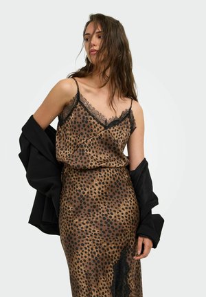 Femme portant une robe nuisette marron et noire à pois avec une bordure en dentelle noire et une veste noire drapée sur ses bras sur un fond uni.