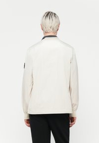 Light beige jacket with a relaxed fit, long sleeves, a ribbed collar, and elastic cuffs. Features a small logo on the left sleeve.