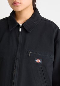 Zwarte zip-up jack van duurzaam materiaal, met een kraag, een voorzak met rits en een klein merklabel op de borst.