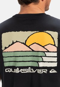 Svart T-skjorte med et fargerikt grafisk design med en gul sol, rosa fjell og grønne bølger, samt "Quiksilver"-tekst nederst.