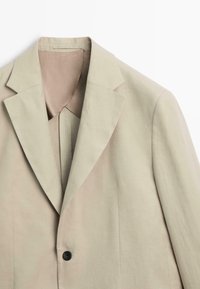 Beige colbert met enkele rij knopen, met schuine revers, één zwarte knoop en een gladde binnenvoering, afgebeeld op een witte achtergrond.