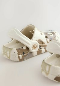 Sabots à motif camouflage blanc avec une sangle arrière, dotés de trous de ventilation et d'un petit emblème d'ours sur la sangle.