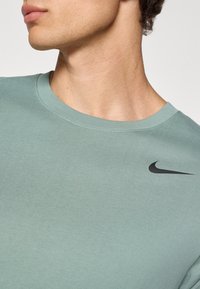 Camiseta de algodón verde claro con cuello redondo y mangas cortas, que presenta un pequeño logo negro de Nike en la parte superior izquierda. Textura suave.