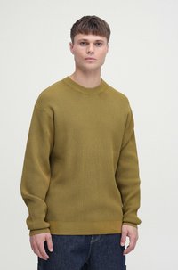 Olive zielony sweter dzianinowy o ribbowanej fakturze, okrągłym dekolcie i długich rękawach, w zestawie z ciemnymi dżinsami. Minimalistyczny design.