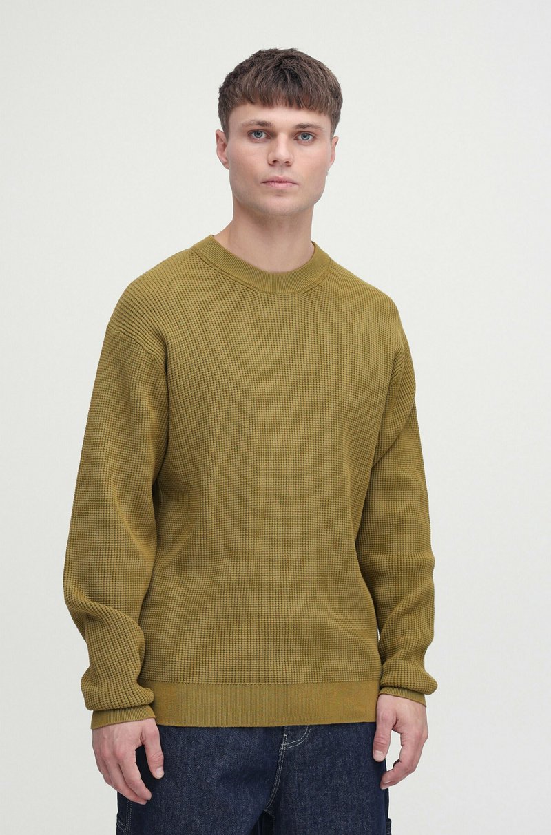 Olive zielony sweter dzianinowy o ribbowanej fakturze, okrągłym dekolcie i długich rękawach, w zestawie z ciemnymi dżinsami. Minimalistyczny design.