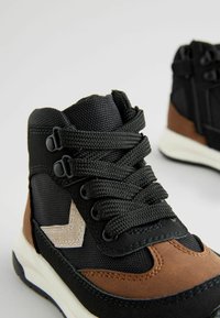 Baskets montantes noires et brunes en matériau textile et cuir, avec un design de lacets texturé et un accent géométrique contrastant.