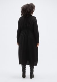 Μαύρο φόρεμα με μακριά μανίκια από υφή υφάσματος, εφαρμοστή μέση και φαρδύ φούστα. Φορεμένο με μαύρες μπότες μέχρι το γόνατο και έχει μανσέτες με κουμπιά.