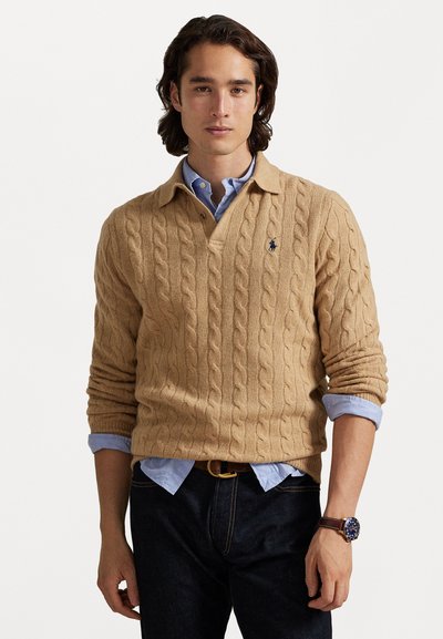 Pull en maille beige avec un col, porté par-dessus une chemise à boutons bleu clair. Un jean foncé et une montre marron complètent la tenue.