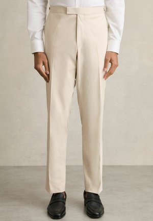 Beige tailored trousers with a flat front, side pockets, and a straight leg design, paired with black loafers. Smooth fabric texture.