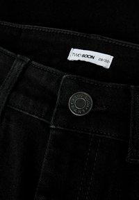 Melnie džinsi ar metāla pogu, uz kuras rakstīts "TWO SOON JEANS", un balto birku ar izmēru 28/32. Izceļas redzama šuvju un tekstūras apdare.