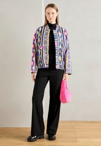 Vrouw in een multicolore, gepatternde jas, zwarte coltrui en broek, met een felroze, fluffy handtas, staand op een houten vloer.