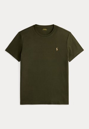 Homme portant un bonnet noir, des lunettes de soleil foncées, un t-shirt vert, un pantalon à motif camouflage et des bottes marron avec des lacets rouges, debout les mains dans les poches.
