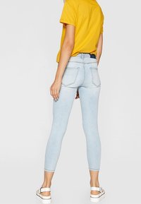 Jeans ajustados de color azul claro, fabricados en denim, con bolsillos traseros y una cintura alta, combinados con una camiseta amarilla y sandalias blancas.