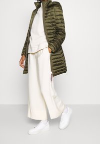 Giacca puffer verde oliva con cappuccio, polsini a costine e chiusura zip, abbinata a una felpa bianca a strati e pantaloni larghi di crema, e sneakers bianche.