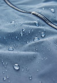 Vandeniui atspari mėlyna medžiaga su lygiu paviršiumi, ant kurio kaupiasi įvairūs vandens lašeliai, ir kontrastingu užtrauktuko akcentu.