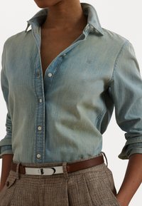 Šviesiai mėlyna džinsinė marškinė su klasikiniu apykakle, mygtukų uždarymu ir sukamu rankovėmis, dėvimi su įstrižais rudais languotais kelnais ir rudomis diržais.