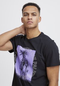 Camiseta de algodón negra con un gráfico púrpura que presenta palmas y texto, cuello redondo, mangas cortas y un corte regular.