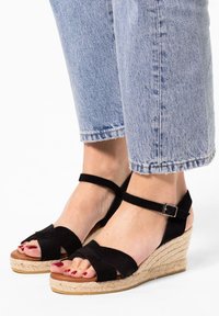Black suede wedge sandals with a braided jute platform, ankle strap with a metal buckle, and crossed straps covering the toes.