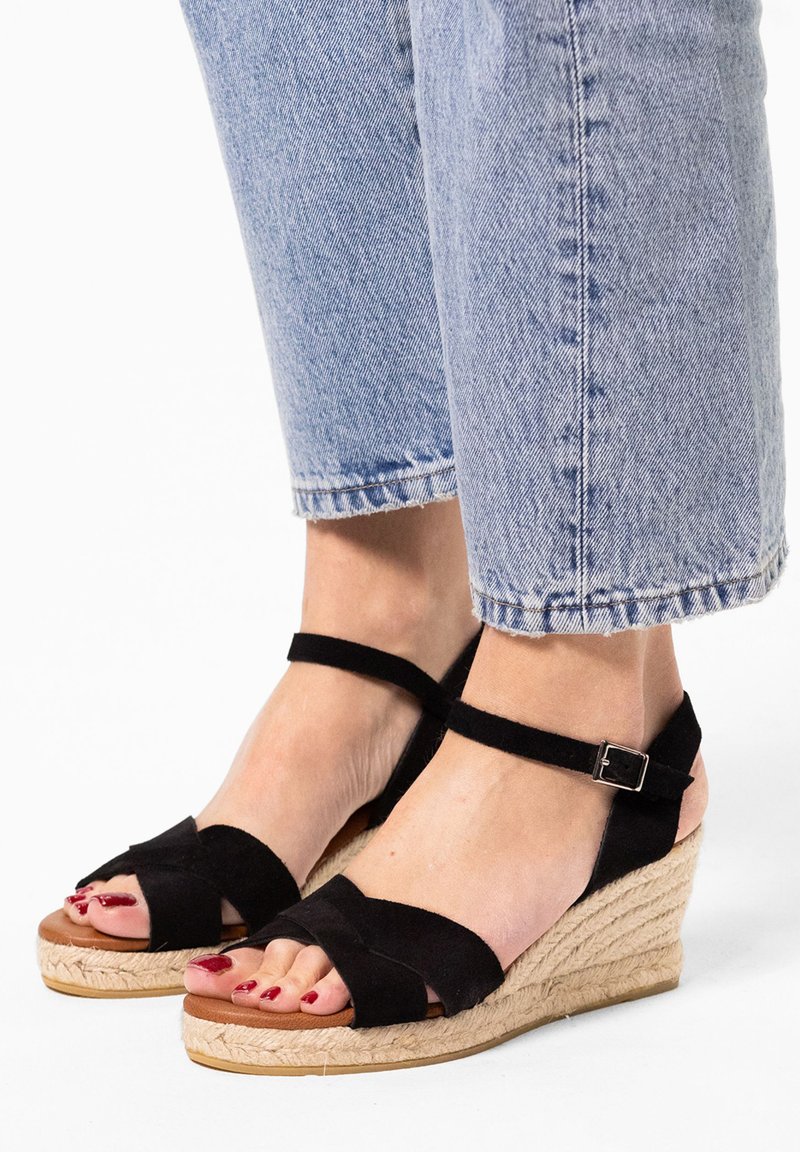 Black suede wedge sandals with a braided jute platform, ankle strap with a metal buckle, and crossed straps covering the toes.