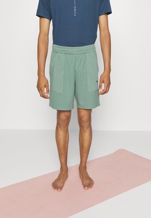 Pantalones cortos verdes acanalados con bolsillos laterales y cintura elástica, combinados con una camiseta azul. De pie sobre una alfombra rosa, con los pies descalzos visibles.