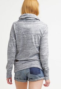 Blondýnka oblečená v šedém dlouhém topu s kruhovým výstřihem a džínových šortkách s tmavým vláknem na pravé zadní kapse, stojící zády.