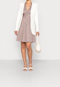 Blazer blanco sobre un vestido marrón con patrón geométrico. Un bolso beige y unos tacones abiertos a juego completan el look.