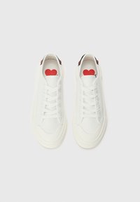 Sneakers brancos com bicos arredondados, couro liso, atacadores e detalhes em preto no calcanhar. O interior apresenta um gráfico de coração vermelho com texto em preto.