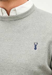 Pull en maille gris avec un col côtelé et un petit logo de cerf bleu sur la poitrine, superposé à une chemise blanche à col.