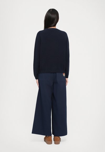 Navy blue sweater with ribbed hem, paired with wide-leg navy trousers featuring two back pockets and a logo patch, worn with brown platform shoes.