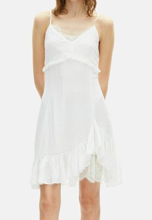 Vrouw in een witte mouwloze jurk met kanten details, ruches aan de zoom en dunne schouderbandjes tegen een effen achtergrond.