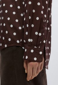 Καφέ πουκάμισο με λευκές πουά, μακριά μανίκια με κουμπωτές μανσέτες και ελαφρώς καμπυλωτό τελείωμα. Ομαλή υφή υφάσματος.