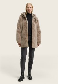 Kunstfelljacke in Hellbraun mit Kapuze, Frontreißverschluss und seitlichen Taschen, kombiniert mit schwarzen Hosen und klobigen schwarzen Stiefeletten.