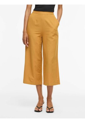 Žena nosi široké krátke nohavice horčicovej farby so stranovými vreckami a čierne sandále, stojaca pred bielym pozadím.