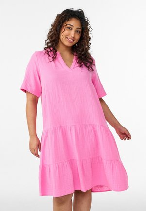 Moteris su garbanotais plaukais dėvi laisvą, kelių ilgio ryškiai rožinę suknelę su trumpomis rankovėmis ir V formos iškirpte, šypsosi į kamerą.