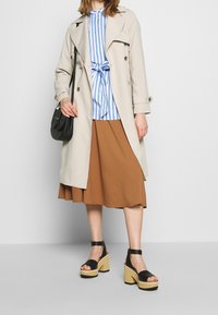 Béžový trenčkot, modro-bílá pruhovaná halenka s uvázaným pasem, hnědá sukně a černé sandály na platformě s páskem kolem kotníku. Černá kabelka.