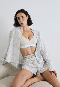 Žena s tamnom kosom do ramena pozira u opuštenom stavu, noseći prugastu majicu i donje rublje Calvin Klein, naslonjena na neutralnu zid.
