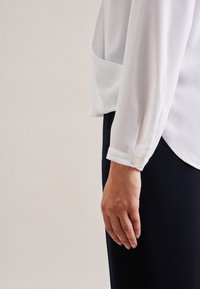 Person, die eine weiße Bluse mit langen Ärmeln und Knopfmanschetten sowie dunklen Hose trägt, steht vor einem einfarbigen hellen Hintergrund.