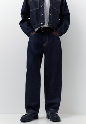 Person wearing dark blue denim jacket and loose-fit jeans with black belt and black leather shoes, hands in pockets.