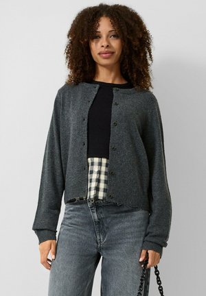 Frau mit lockigem Haar steht und trägt eine dunkelgraue, geknöpfte Strickjacke, ein schwarzes Top, ein kariertes Hemd, graue Jeans und hält eine schwarze Tasche mit Kettenriemen.