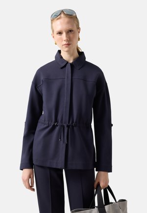 Femme portant une veste bleu marine avec taille à cordon, pantalon assorti, boucles d'oreilles en or en forme de créoles, lunettes de soleil sur la tête et tenant un sac fourre-tout gris.