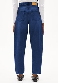 Blauwe high-waisted jeans met een ontspannen pasvorm, zichtbare stiksels en twee achterzakken. Gecombineerd met zwarte schoenen met zilveren accenten.