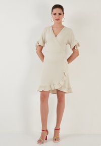 Light beige wrap dress with short sleeves, ruffled hem, subtle texture, and tied accents at the sleeves. Paired with multicolored strappy heels.